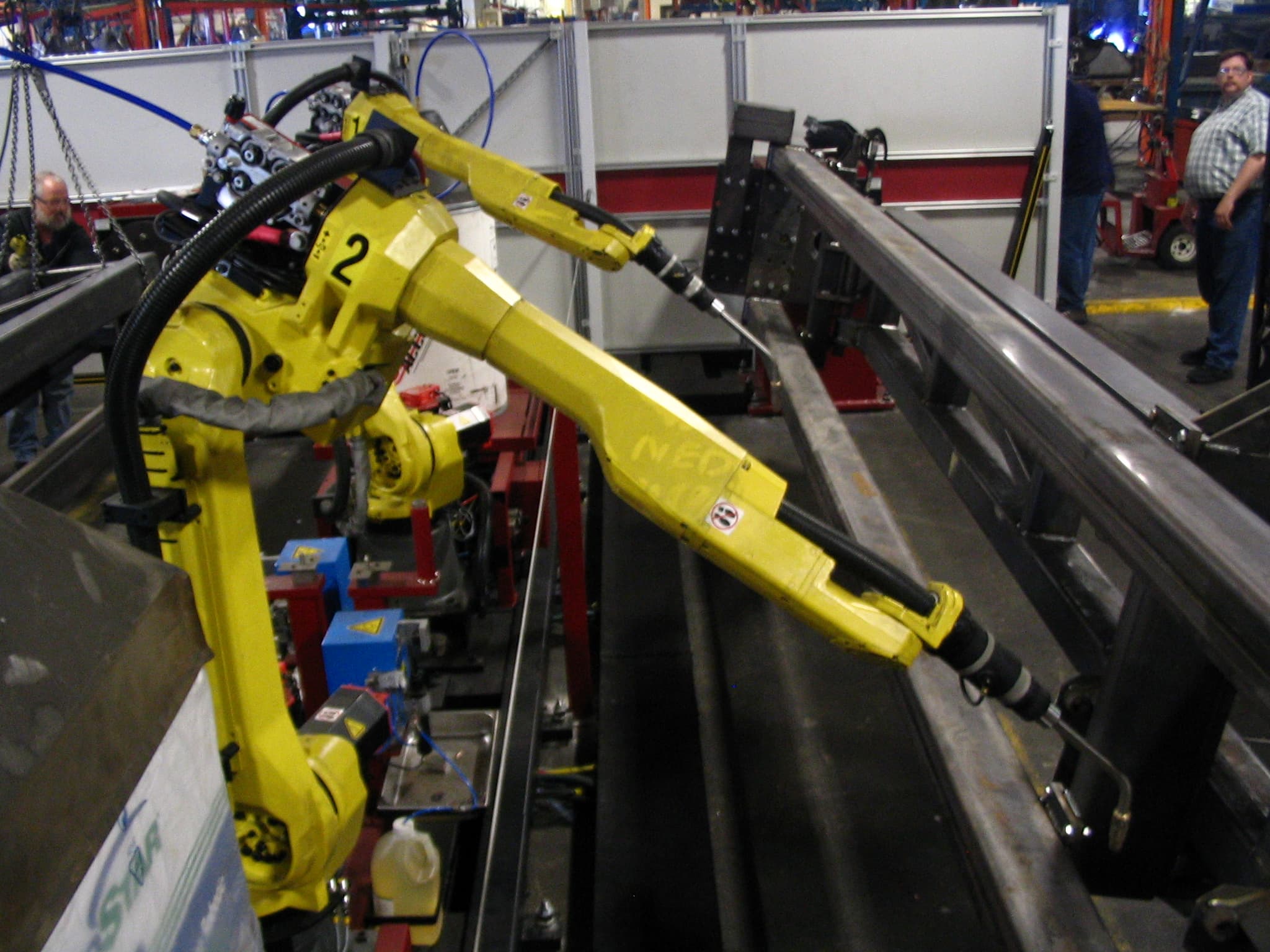 Two FANUC six-axis industrial welding robots on a factory floor, orange-liveried arms raised and preparing to weld a part fixtured on the line in front of them. The image is used here because Anthropic's September 30, 2026 paper What work can robots do? defines its highest-capability tier, E1, as the environment where robots can perform work today in purpose-built facilities such as manufacturing lines like this one — the condition under which 23% of U.S. working hours fall exposed. The paper argues that stepping out of this kind of purpose-built environment into warehouses (E2) adds only another 10% of working hours, and into unstructured settings like city streets (E3) only another 1%, which is where the economic case for robotics mostly already is.
