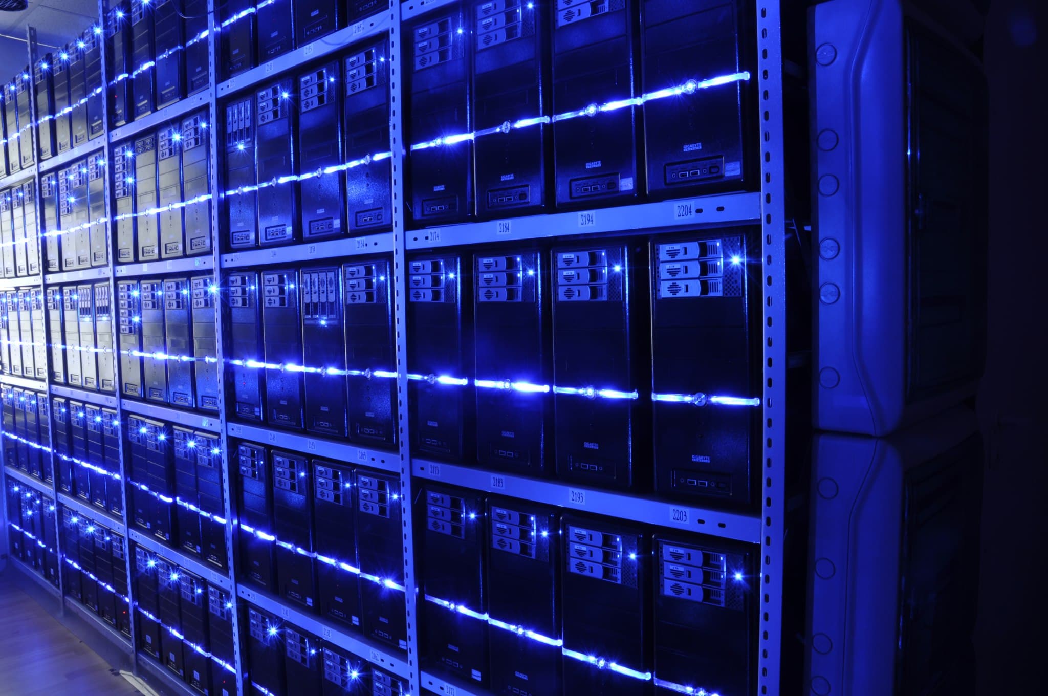 A wide interior view of a modern data center, showing two long parallel rows of black server racks running to a vanishing point under blue-lit overhead lighting, with a raised metal floor and cabling trays overhead. The image stands in for Oracle's Project Jupiter, a 2.45-gigawatt Stargate campus under construction in Doña Ana County, New Mexico, on which Oracle served developer Blue Owl Capital a force majeure notice on September 24, 2026, citing energy-infrastructure delays that put the 2028 completion date at risk.
