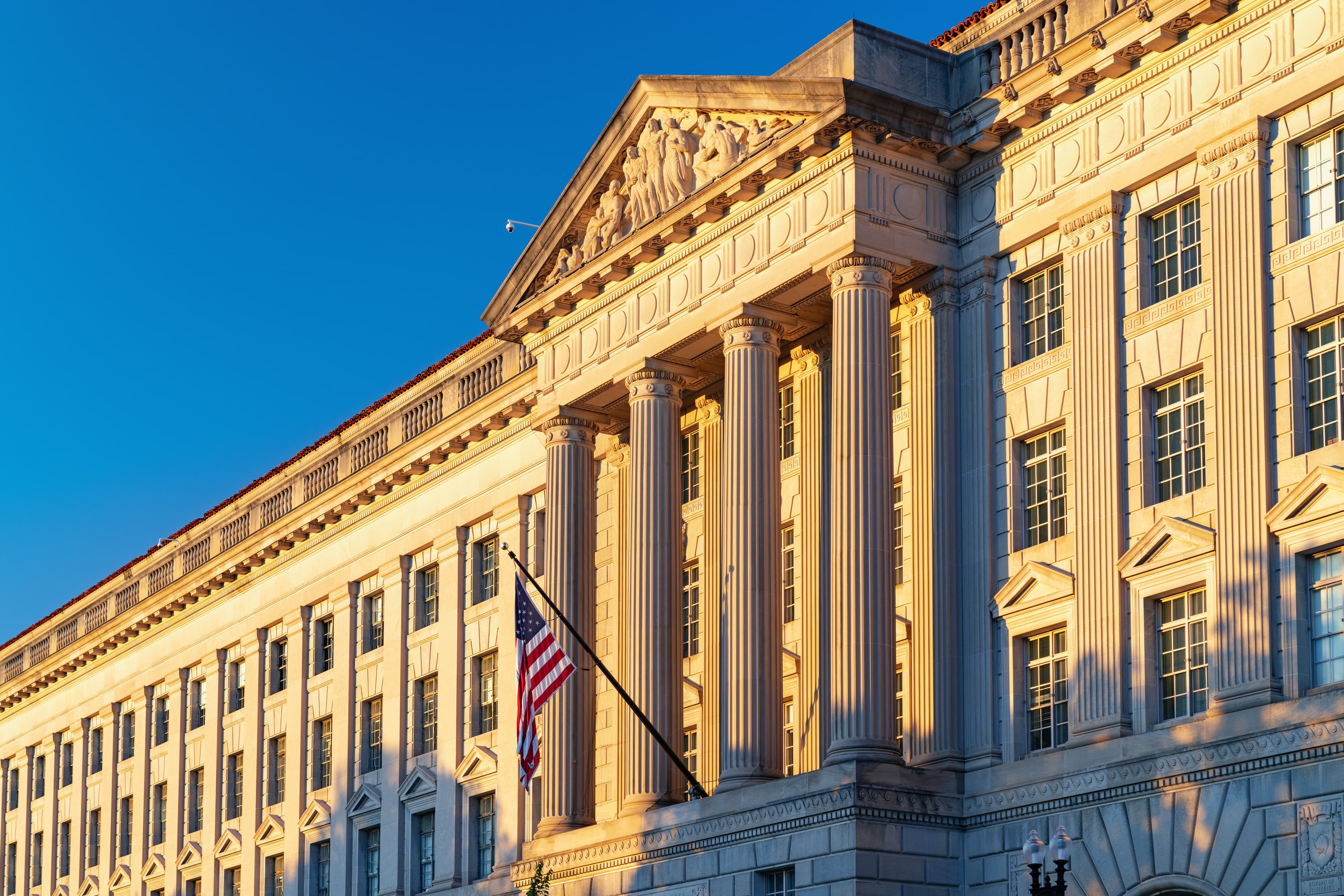 A colour photograph of the Herbert C. Hoover Federal Building at 1401 Constitution Avenue in Washington, D.C., headquarters of the U.S. Department of Commerce. The neoclassical limestone facade with its columns rises under a clear sky, seen from the street. On Tuesday June 30, 2026, Commerce Secretary Howard Lutnick sent Anthropic a letter from this building notifying the company that the export controls applied to Claude Fable 5 and Mythos 5 on June 12 had been lifted after nineteen days offline, replacing the export-license requirement with a set of standing obligations covering security-risk detection, release-protocol coordination, and malicious-activity reporting.