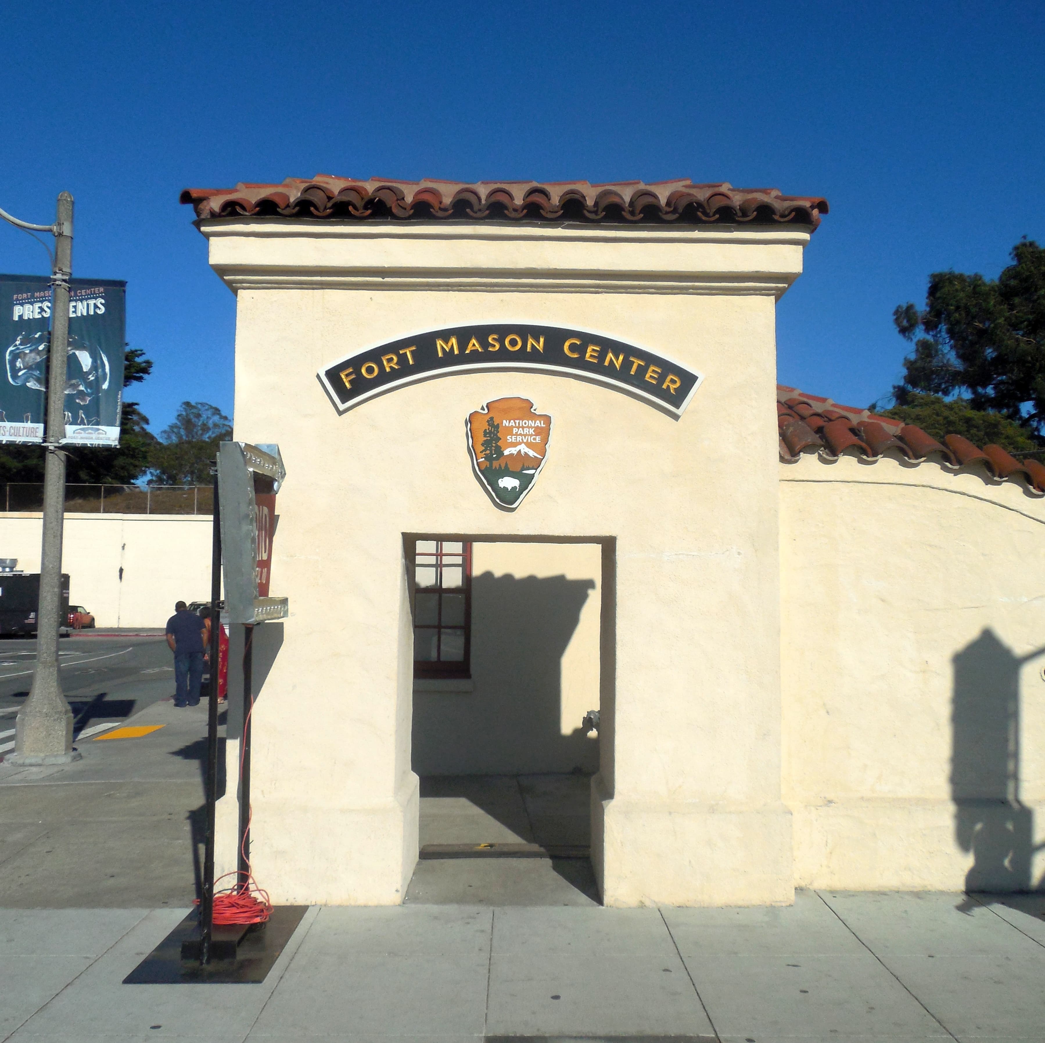 A daytime colour photograph of the western entrance to Fort Mason Center in San Francisco, taken near the Farmers' Market on September 18, 2015 — a low square stone gate marker reading "Fort Mason Center" set on a paved corner, with an access road, scattered trees, and the pale roofs of the Center's old Army warehouses visible beyond the entrance. Fort Mason Center is the historic former US Army post on San Francisco's northern waterfront, now a non-profit cultural campus run by the Fort Mason Center for Arts and Culture, and it was the venue of Cursor's inaugural Compile conference on June 16, 2026 — the one-day event at which the company announced its first self-trained 1.5-trillion-parameter foundation model, the GitHub-rival Git platform Origin, and a Cursor Mobile iOS beta, the same day SpaceX disclosed a $60-billion all-stock acquisition of Cursor's parent company Anysphere.