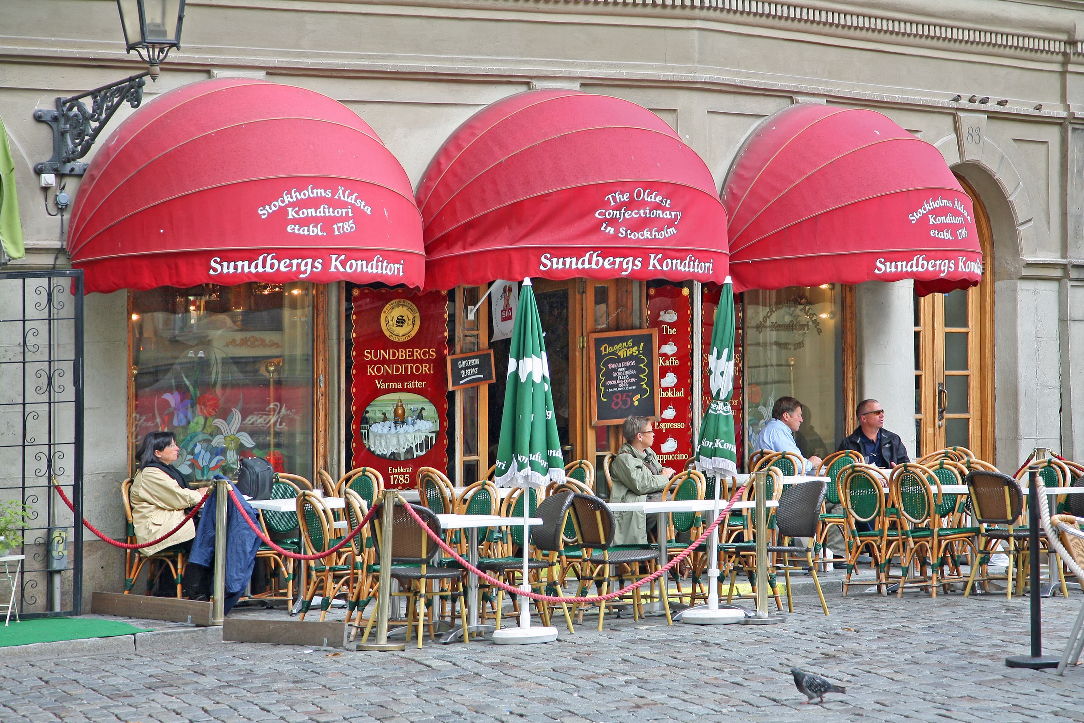 A wood-fronted Stockholm cafe with hanging lamps inside the window and a small handwritten chalkboard on the pavement.
