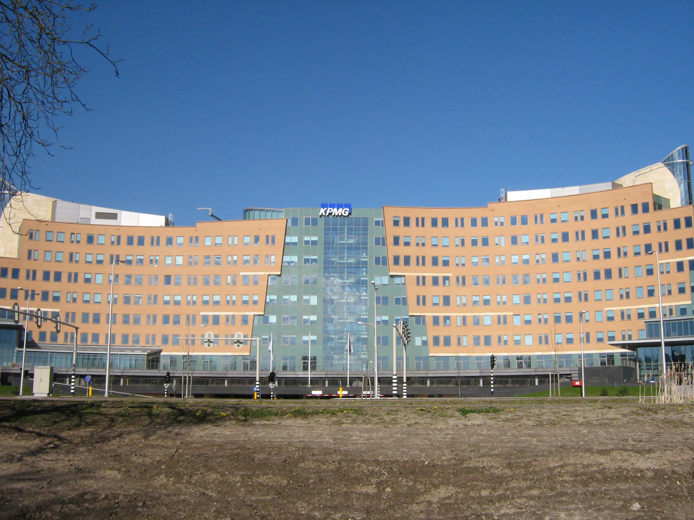 The KPMG global headquarters at 1 Laan van Langerhuize, Amstelveen, in the Netherlands — a large modern corporate complex of stepped glass-and-steel blocks set behind a long reflecting pond and a flagpole flying the KPMG blue ensign. The building has been KPMG International's head office for the global Big Four accountancy and consultancy network since the 2000s. On June 12, 2026 the firm withdrew its flagship report "Redefining excellence in the age of agentic AI" after an audit by detection company GPTZero found that only five of the report's forty-five citations correctly matched their sources.
