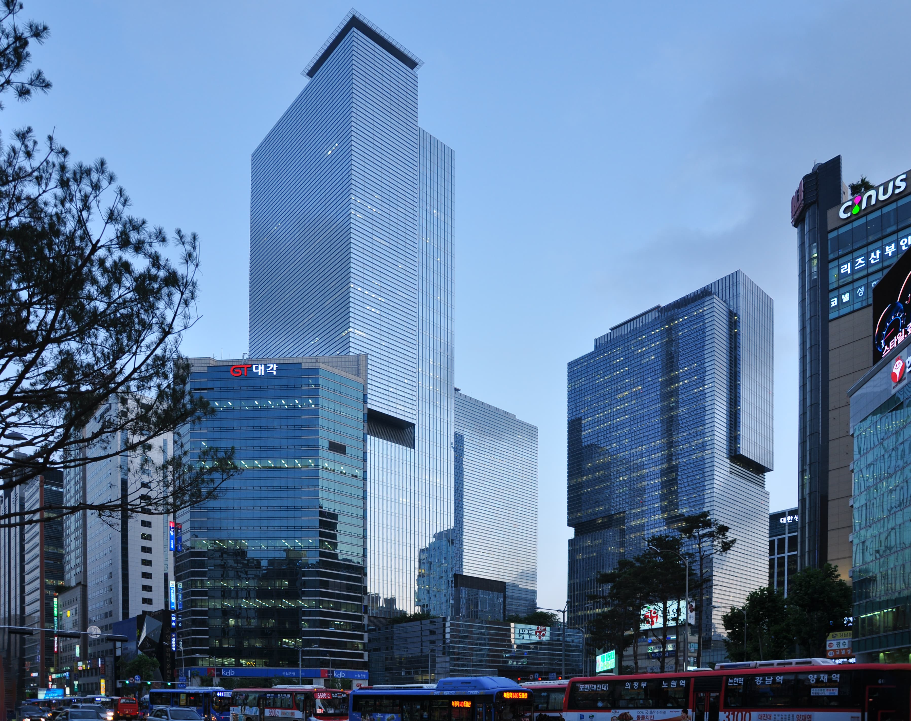 A daytime photograph of the Samsung Town headquarters complex in Seoul's Seocho District — three tall blue-glass towers rising side by side against a clear sky, with the central tower bearing the Samsung wordmark in white near the top, set in front of a wide multi-lane plaza and low landscaped traffic divider. Samsung Town houses Samsung Electronics' executive offices including those of Executive Chairman Lee Jae-yong, who posed at the same Seocho complex with OpenAI CEO Sam Altman on October 1, 2025, after the four-affiliate letter-of-intent that pulled Samsung into the $500 billion Stargate project. On June 22, 2026 the same OpenAI–Samsung relationship was named in the other direction — the company supplying the high-bandwidth DRAM for Stargate's data centres is now the customer rolling ChatGPT Enterprise and Codex out to every Korean employee and every Device eXperience division worker worldwide, in what both companies describe as one of OpenAI's largest enterprise deployments to date.