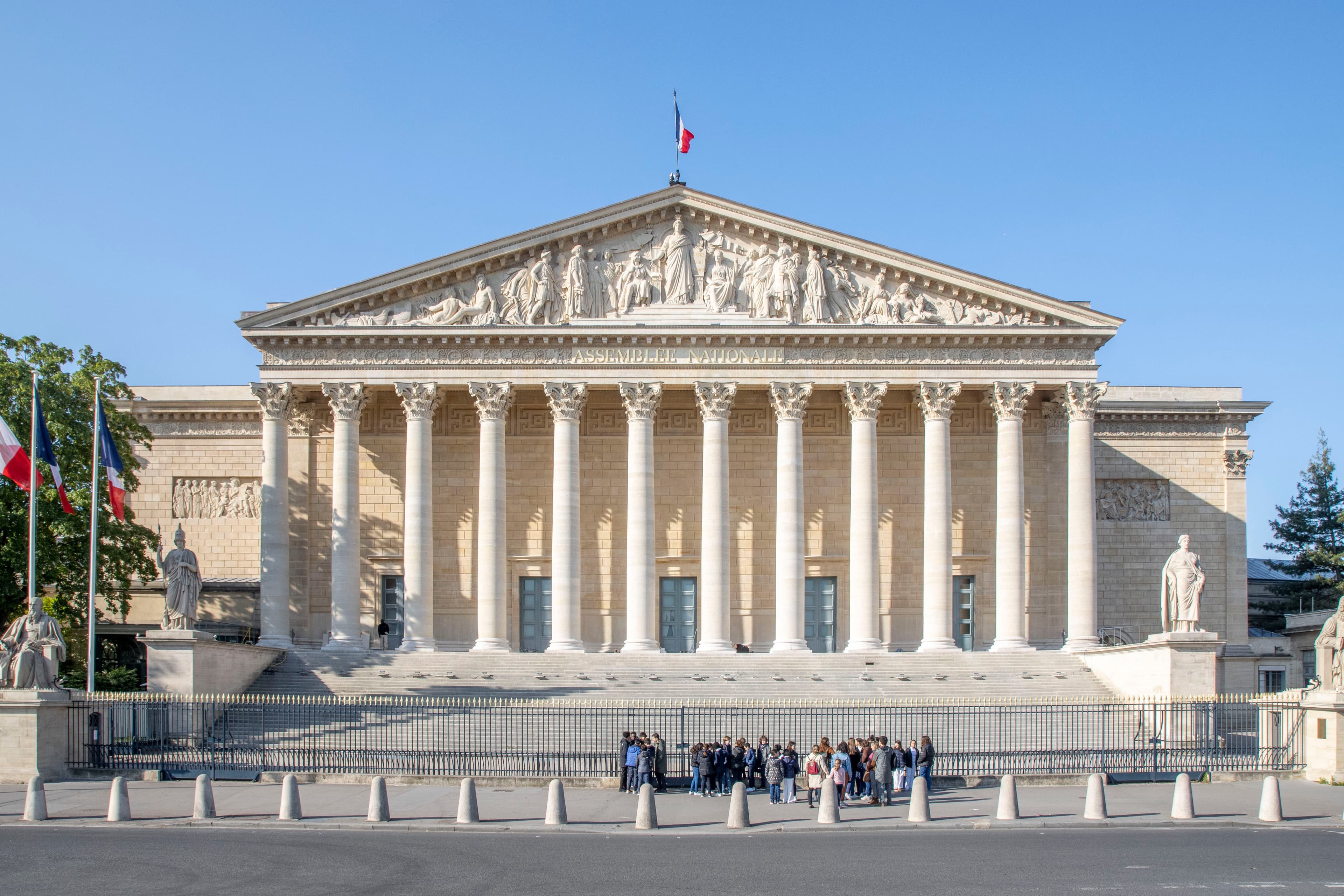 A daytime photo of the north façade of the Palais Bourbon in Paris, the seat of the French National Assembly, showing the colonnaded portico and the tricolour flag flying above the building.