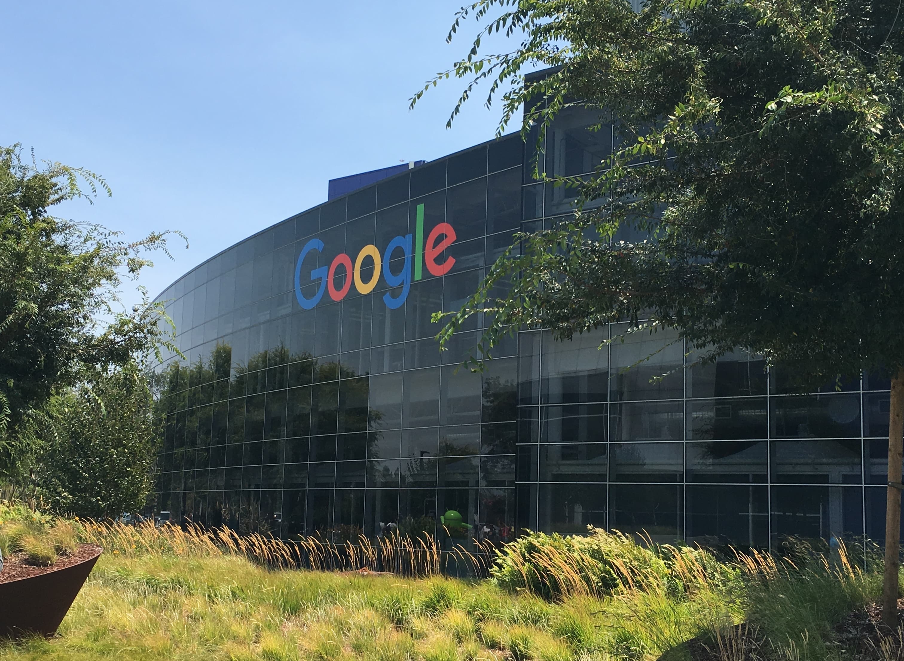 A daytime photograph of the Googleplex, Google's headquarters in Mountain View, California — the colourful Android-and-Google-logo signage in front of a wide glass-and-steel office building under a clear blue sky, with a manicured lawn and palm trees in the foreground. On June 17, 2026, Google's developer blog and Microsoft's commandline blog jointly published the Agentic Resource Discovery (ARD) specification — an open standard for how AI agents discover tools, MCP servers, OpenAPI APIs, and other agents on the open web. Launch contributors named on agenticresourcediscovery.org were Cisco, Databricks, GitHub, GoDaddy, Google, Hugging Face, Microsoft, Nvidia, Salesforce, ServiceNow, and Snowflake.