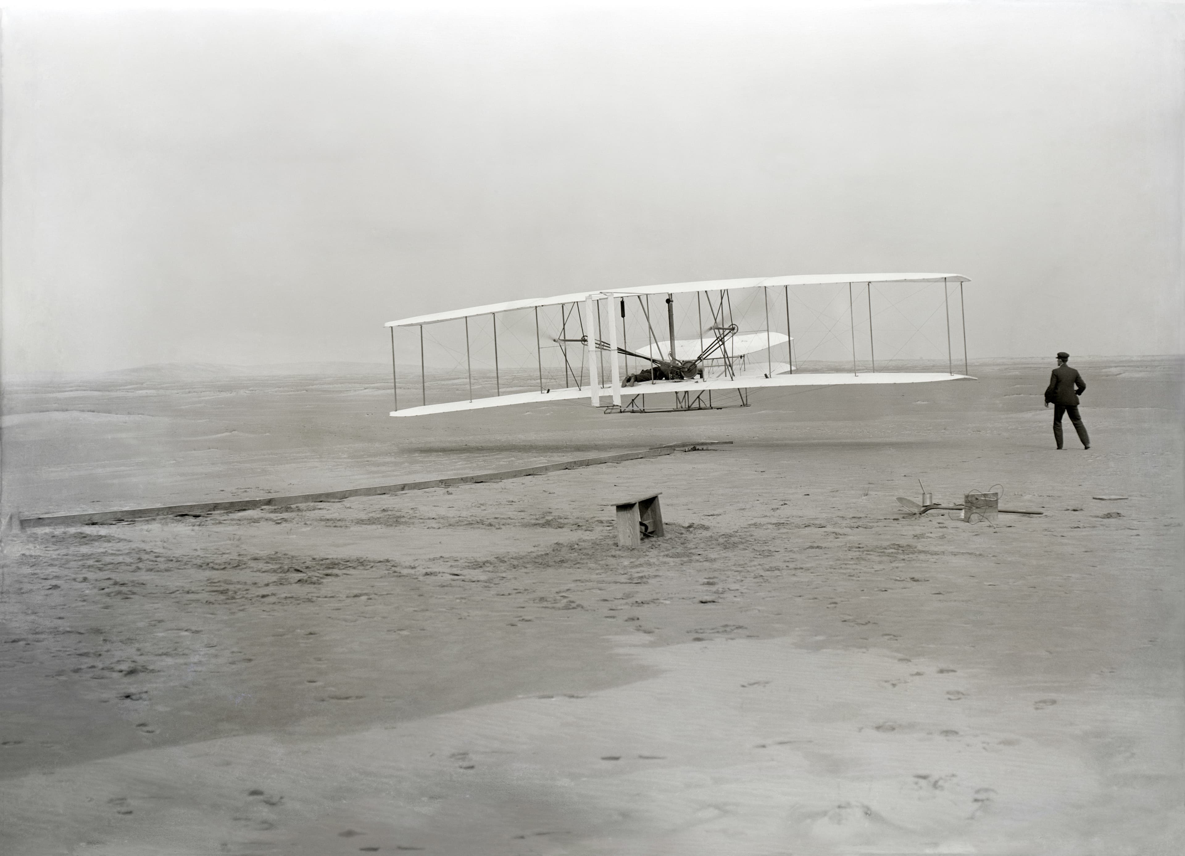 Black-and-white photograph from December 17, 1903 of the Wright Flyer taking off from Kitty Hawk, North Carolina. Orville Wright is at the controls; Wilbur Wright runs alongside the right wingtip. The aircraft is a few feet off the sand.