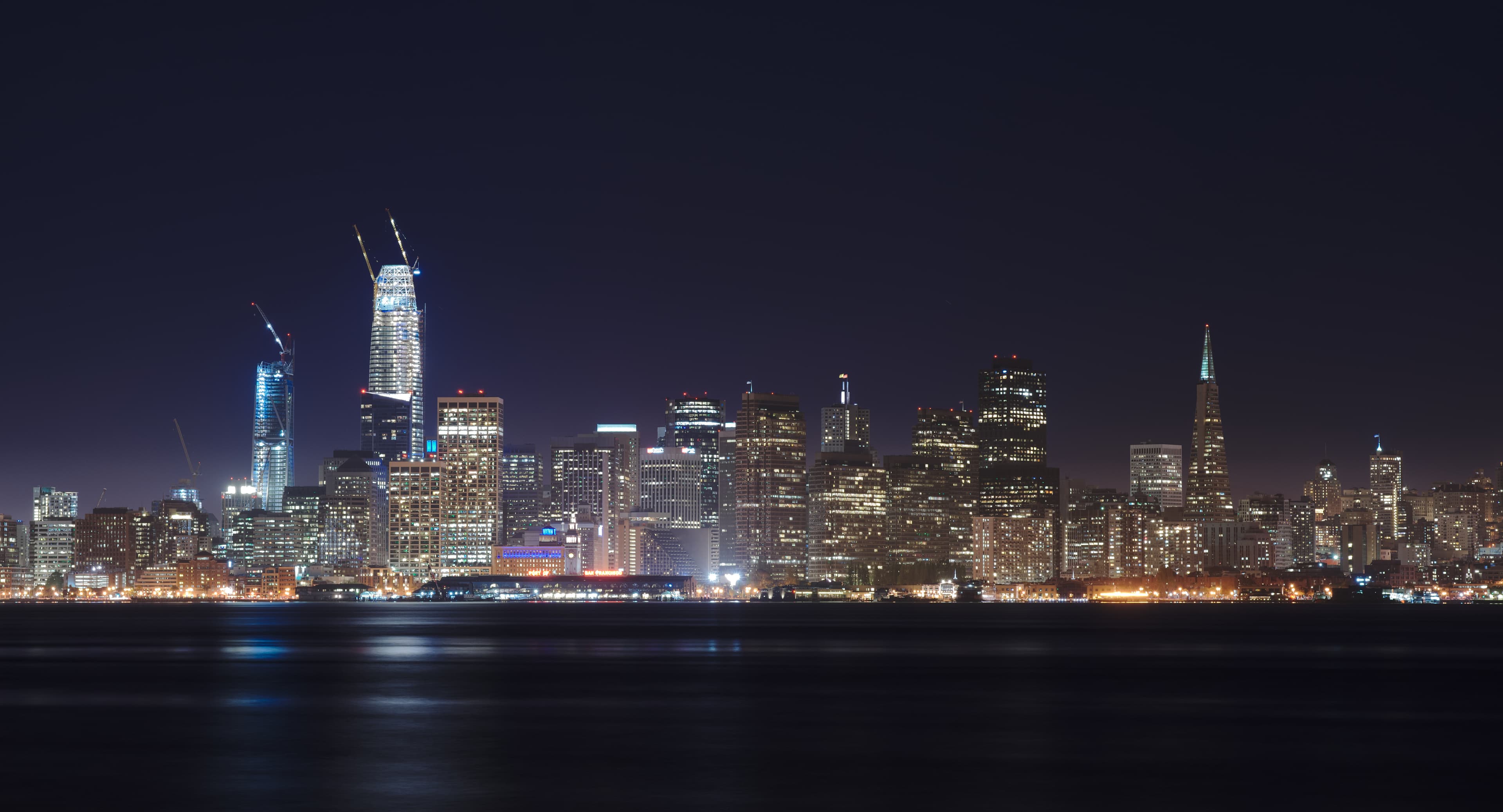 A wide panorama of the San Francisco downtown skyline at dusk, viewed from Treasure Island, with the Bay Bridge curving toward the city and the Salesforce Tower lit up against the sky.