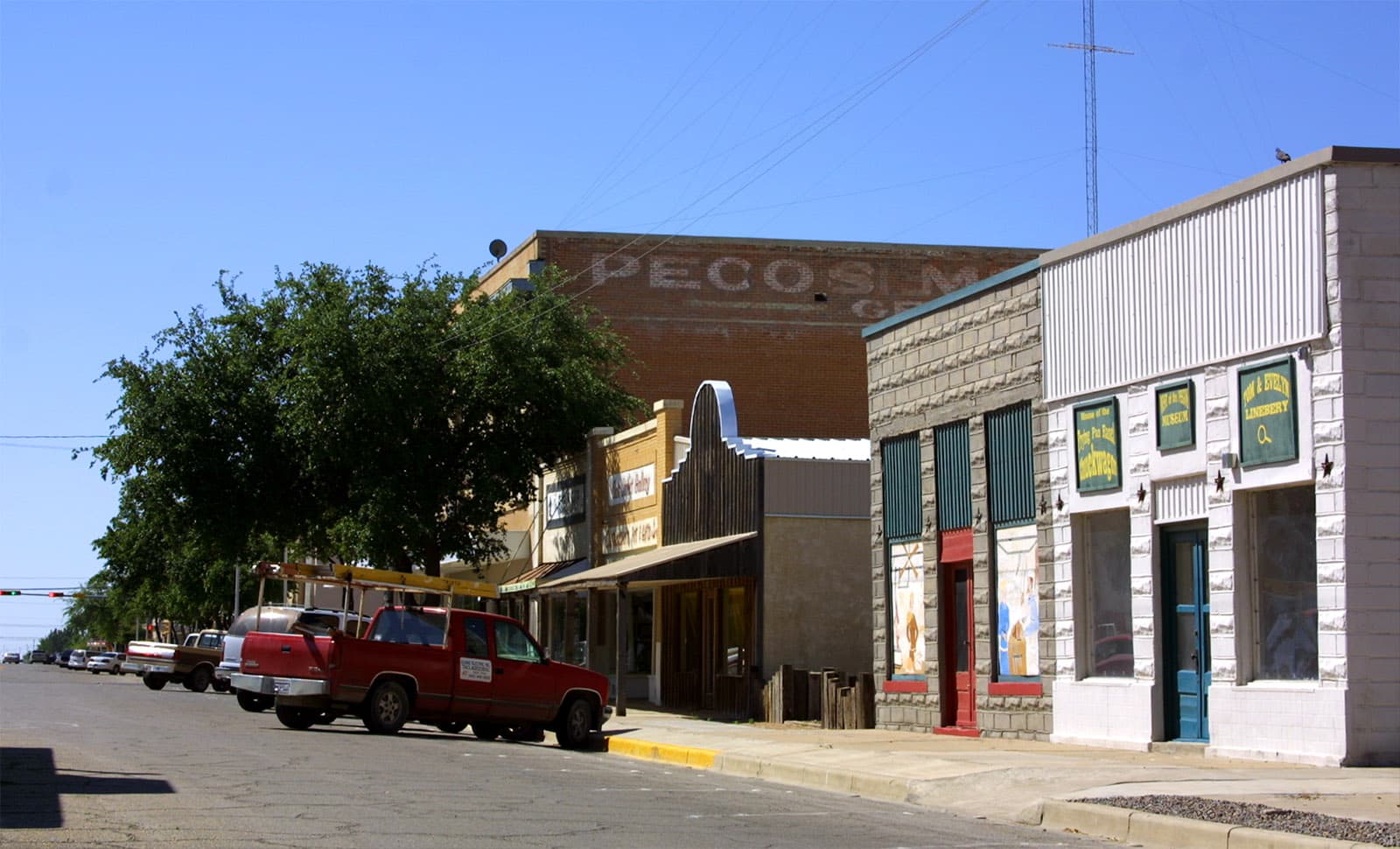 Storefronts in downtown Pecos, Texas, photographed in April 2004 — low single-storey buildings along a wide West Texas main street in a town of about thirteen thousand people in Reeves County, on the edge of the Permian Basin. Pecos is the announced site of Microsoft's 2-gigawatt data centre campus, fed by a 20-year behind-the-meter natural gas plant developed with Chevron, Engine No. 1, and GE Vernova, scaling to 2.67 GW of generation by 2028. Pecos also claims to be the venue of the world's first rodeo, held July 4, 1883.