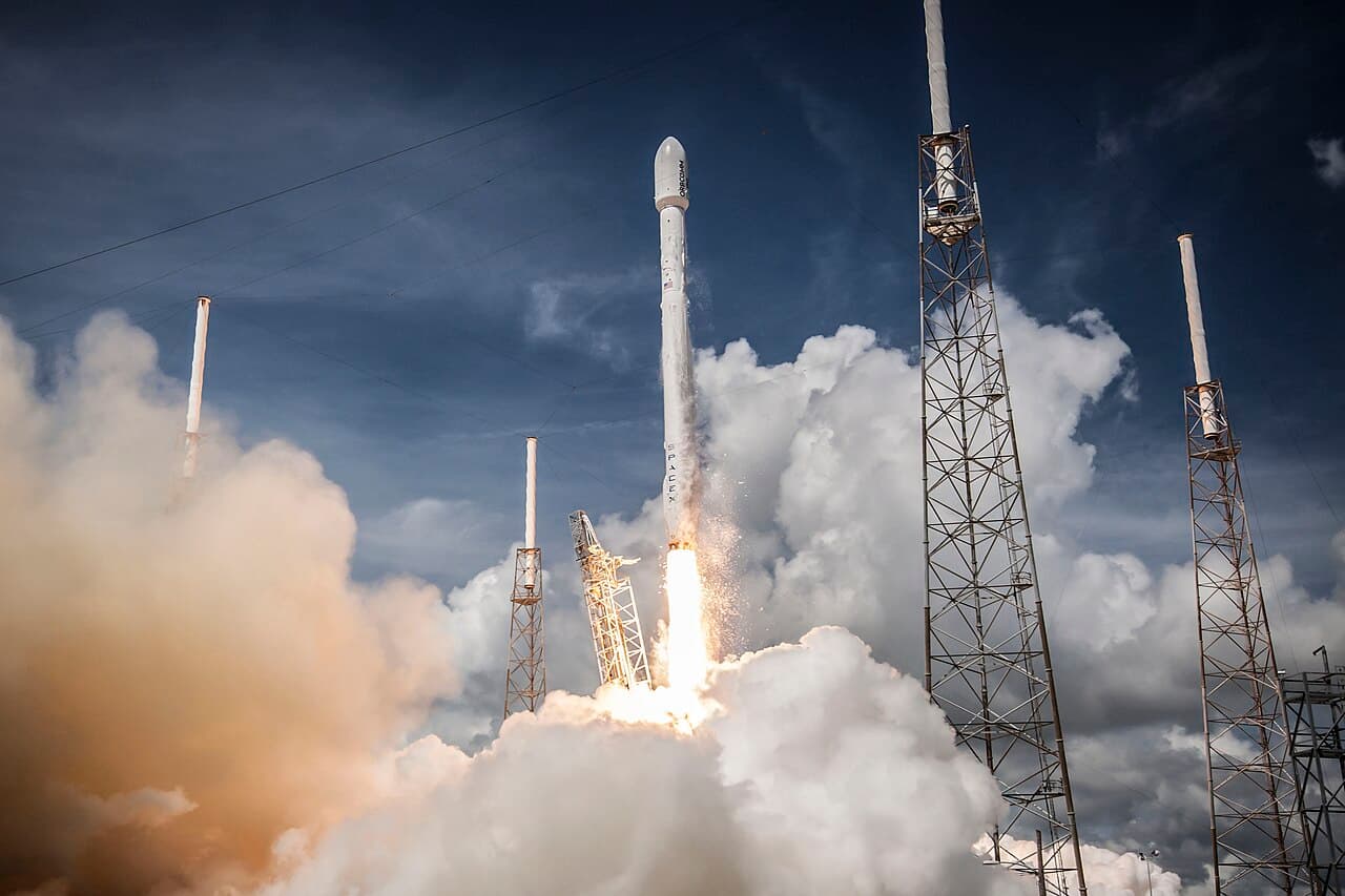 A SpaceX Falcon 9 rocket lifting off at night, the first-stage exhaust illuminating the launch tower.
