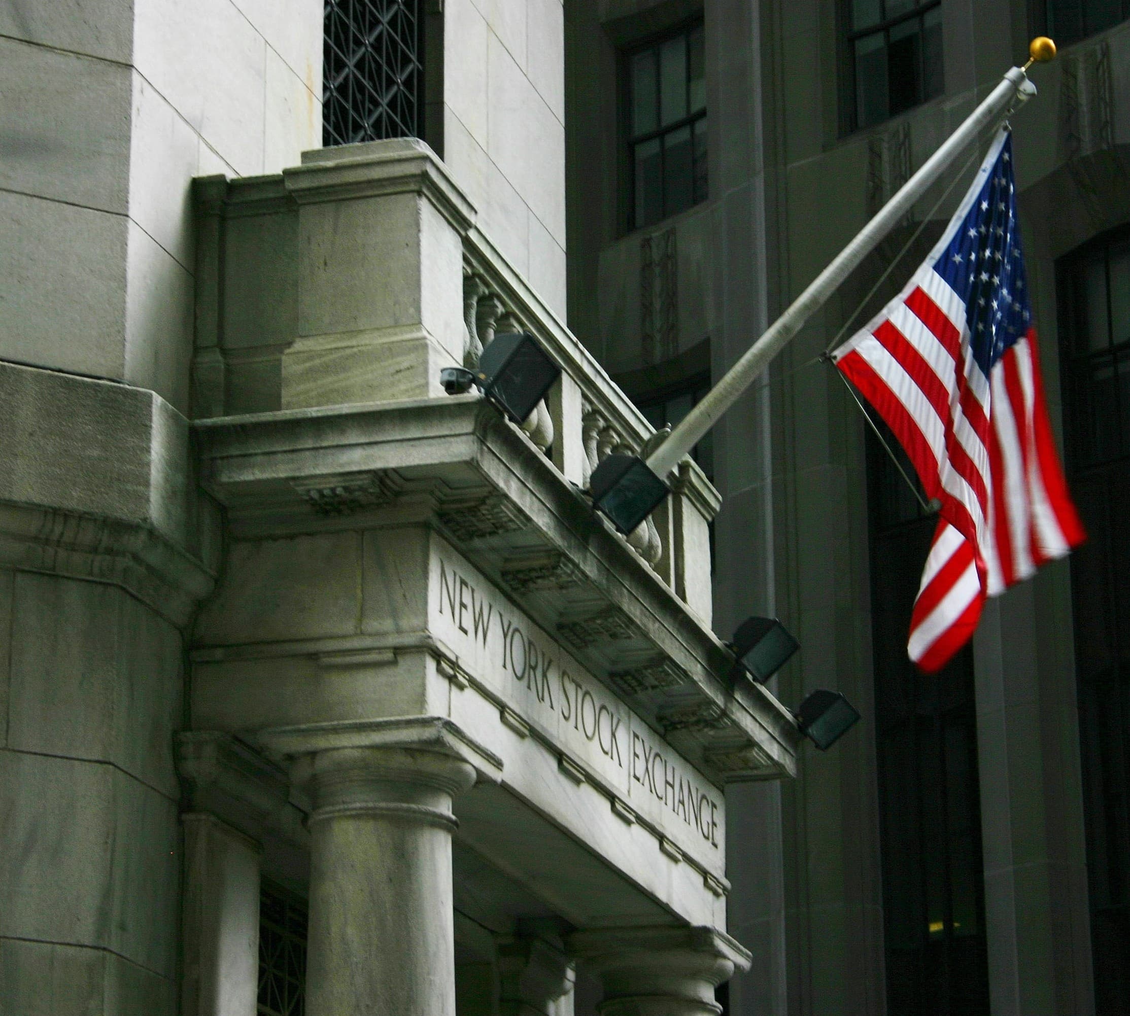 The neoclassical marble facade of the New York Stock Exchange on Wall Street, with Corinthian columns and the giant American flag draped across the front.